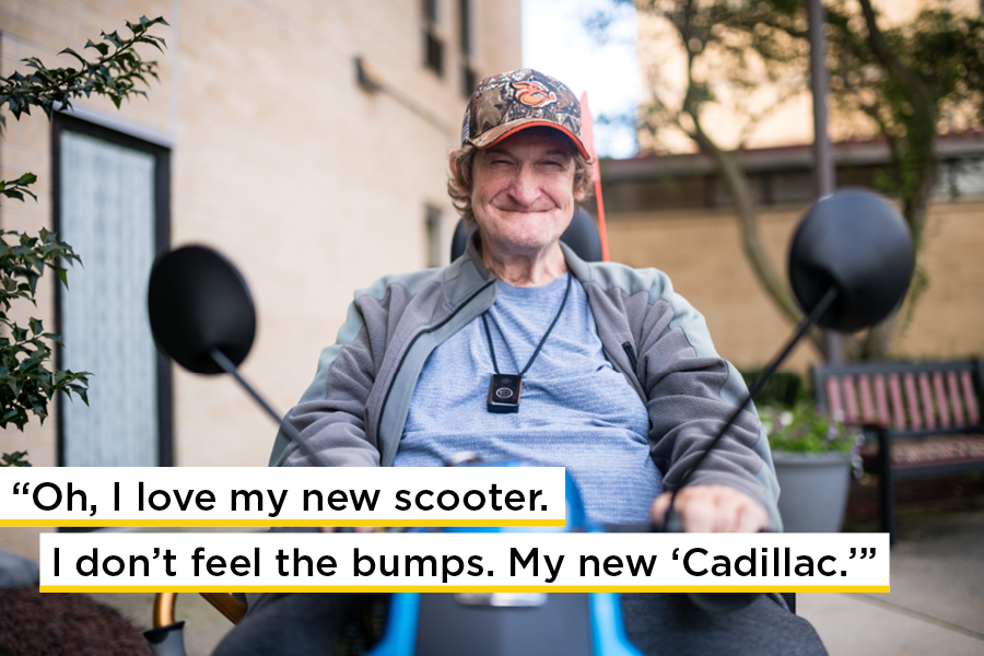 An older white man smiles seated in his blue mobility scooter outside in front of a bench and an apartment building. A quote is written across the photo that says Oh I love my new scooter. I dont feel the bumps. My new Cadillac.
